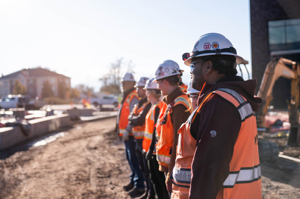 Murray City Hall Project A group of construction workers stand in a line at a construction site, wearing hard hats and high-visibility vests. Bright sunlight illuminates the scene.